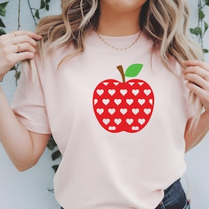 Op de afbeelding: Een lichtroze t-shirt met een groot rood appelontwerp gevuld met witte harten. De appel heeft een bruine steel en een groen blad. Het shirt is een casual stijl.