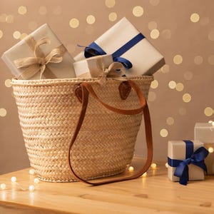 Image of a Straw Basket Bag with long and short chestnut leather handles. Basket is on a table and is filled with wrapped gifts, surrounded by fairy lights.
