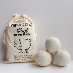 Peut inclure: Ensemble de quatre boules de séchage en laine blanche dans un sac en coton blanc à cordon avec du texte noir qui dit "Sand & Salt Wool Dryer Balls" et un mouton de dessin animé. Le texte sous le mouton dit "4 Extra Large Dryer Balls 100% New Zealand Wool www.sandandsalt.co.uk"