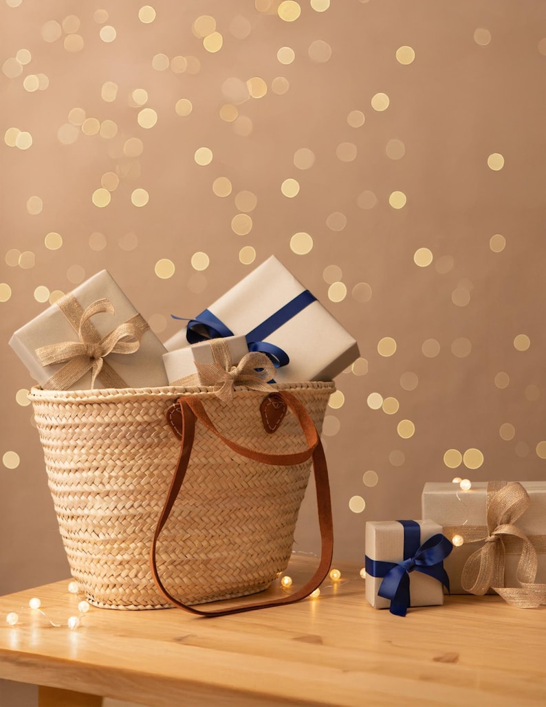 Image of a Straw Basket Bag with long and short chestnut leather handles. Basket is on a table and is filled with wrapped gifts, surrounded by fairy lights.
