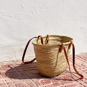 Image of a Straw Basket Bag with long and short chestnut leather handles. Basket is on a terracotta tiled floor against a whitewashed wall in the sunshine.