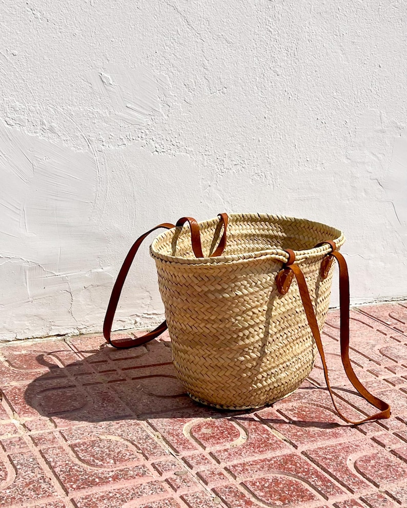 Image of a Straw Basket Bag with long and short chestnut leather handles. Basket is on a terracotta tiled floor against a whitewashed wall in the sunshine.