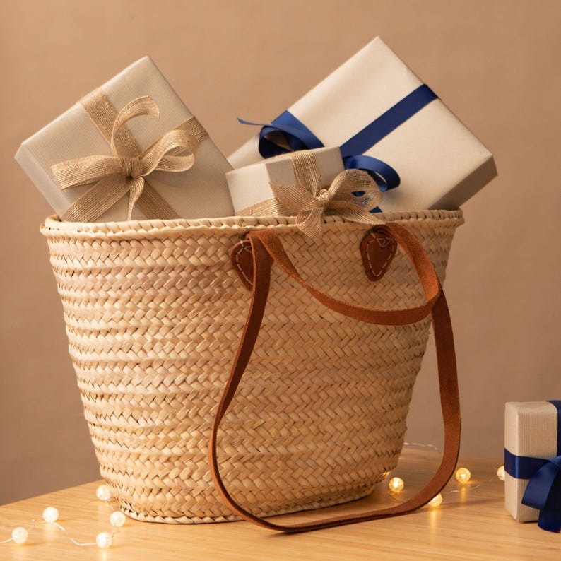 Peut inclure: Un panier en osier avec des anses en cuir marron est rempli de cadeaux emball&eacute;s. Les cadeaux sont envelopp&eacute;s dans du papier beige et attach&eacute;s avec des rubans beiges et bleus. Le panier est pos&eacute; sur une surface en bois avec des guirlandes lumineuses.