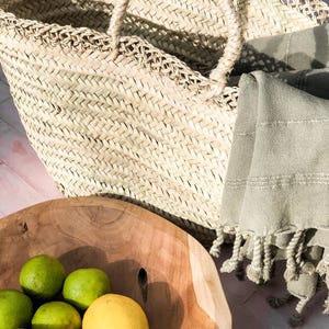 Peut inclure: Un bol en bois rempli de citrons et de limes. Le bol est posé sur une surface rose à côté d'un sac en paille tressé et d'une serviette verte.