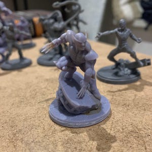 May include: A light purple 3D printed miniature figurine of a character in a crouching pose with extended claws on both hands, mounted on a circular base. The figure features pointed headgear and shoulder pads, rendered in a solid monochromatic resin.