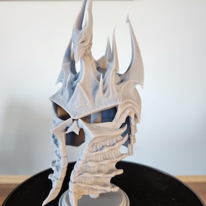 Peut inclure: Casque de fantaisie gris clair avec des détails complexes, notamment de longues cornes pointues et une surface texturée. Le casque est monté sur une base circulaire. Le design suggère un personnage de jeu vidéo ou de film fantastique.