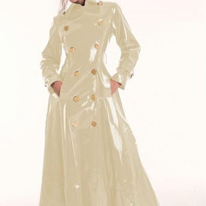 Könnte beinhalten: Ein langer, cremefarbener, zweireihiger Trenchcoat aus einem glänzenden, lackartigen Material. Der Mantel verfügt über einen breiten Kragen, lange Ärmel mit Manschetten, zwei Seitentaschen und zwei vertikale Reihen runder, goldfarbener Knöpfe.