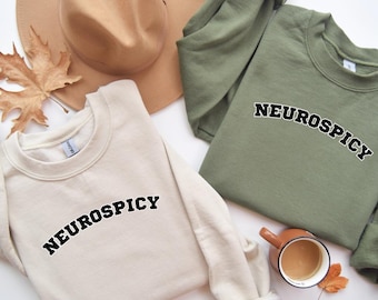 May include: Two sweatshirts with the text "Neurospicy" printed on them in a black, arched font. One sweatshirt is cream colored and the other is olive green.