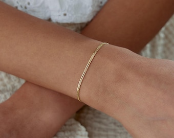 Könnte beinhalten: Ein dünnes, zartes Goldkettenarmband mit kleinen, ineinandergreifenden Gliedern. Das Armband wird um das Handgelenk getragen und weist eine schlichte, polierte Metalloberfläche auf, die das Licht auf ihrer gesamten Oberfläche reflektiert.