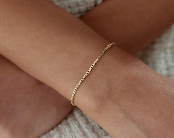 Könnte beinhalten: Ein schmales Armband aus einer goldenen Boxkette, die aus einer fortlaufenden Reihe kleiner, quadratischer Glieder besteht. Die gelbgoldene Kette liegt auf der Haut und zeichnet sich durch eine gleichmäßige Textur und eine glatte Oberfläche aus.