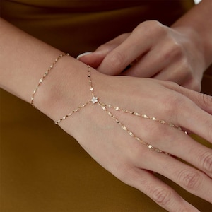 Könnte beinhalten: Ein zartes Handkettchen aus Gold mit einer dünnen, schimmernden Kette, die um das Handgelenk verläuft und mit einer Fingerschlaufe verbunden ist. Ein kleiner, blumenförmiger Anhänger mit winzigen klaren Steinen sitzt in der Handmitte.