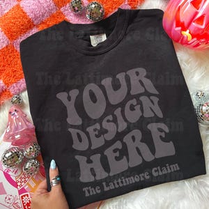 May include: Black t-shirt with the text "YOUR DESIGN HERE" in a retro font. The text "The Lattimore Claim" is below the text. The shirt is on a white surface with decorative items.