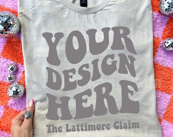May include: A light beige t-shirt with the text "YOUR DESIGN HERE" in gray, with "The Lattimore Claim" below. The shirt is surrounded by small disco balls on a pink and orange checkered surface.