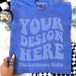 May include: A periwinkle blue t-shirt with the words "YOUR DESIGN HERE" in a light blue, retro font. The text "The Lattimore Claim" is below. The shirt is on a white fluffy surface with disco balls and a black and white checkered pattern.