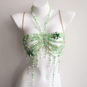 May include: A green and white beaded top with floral details and dangling beads, displayed on a white mannequin. The top has gold chain straps and a halter neck design. The intricate pattern is layered, creating a unique aesthetic.
