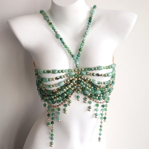 Op de afbeelding: Een groene kralen body chain met een halter-stijl halslijn en meerdere horizontale strengen over de borst. Het sieraad heeft verschillende groentinten en goudkleurige accenten, met bungelende kralen strengen.