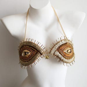 Könnte beinhalten: Ein goldfarbenes Neckholder-Oberteil mit zwei augenförmigen Cups, die mit aufwendigen Perlenarbeiten, metallischen Pailletten und strahlenförmigen Stacheln verziert sind. Die Mitte ist durch einen kleinen sonnenförmigen Anhänger verbunden.