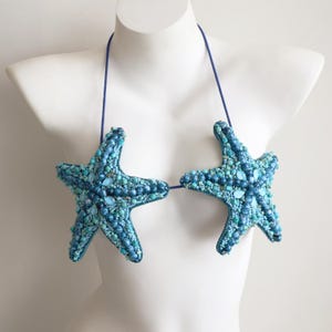 Può includere: Un top bikini all'americana con due coppe a forma di stella marina, ricoperte da una fitta disposizione di perline, pietre e cristalli blu e turchesi. Il capo è tenuto insieme da sottili spalline blu che partono dalla parte superiore.