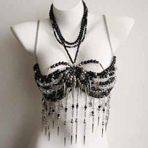 May include: A decorative body harness featuring black and clear faceted beads, silver-toned metal chains, and sharp conical spikes. The piece includes a multi-strand beaded necklace section, a structured bodice area, and dangling fringe accents.