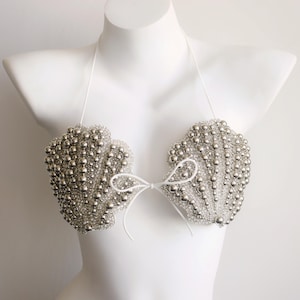 Könnte beinhalten: Ein Neckholder-Bikini-Oberteil mit zwei muschelförmigen Cups, die mit silbernen, metallischen Perlen in verschiedenen Größen besetzt sind. Das Kleidungsstück verfügt über dünne weiße Bänder am Nacken und in der Mitte, um es zu befestigen.