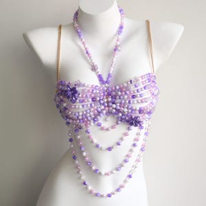 May include: A beaded bustier in shades of purple, lavender, and white, displayed on a white mannequin. The bustier features multiple strands of beads, with floral accents and gold chain straps. The design includes a necklace and cascading beaded strands.