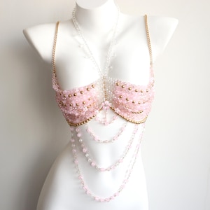 May include: A pink beaded bustier with gold chain straps and accents. The bustier features multiple strands of pink and clear beads cascading down the front. The garment is displayed on a white mannequin.