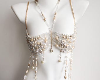May include: A decorative body harness featuring a structured bra-like bodice crafted from clear beads, white pearls, and tan stone-like accents. The piece includes gold-tone chain straps and dangling beaded fringe elements that hang from the base.