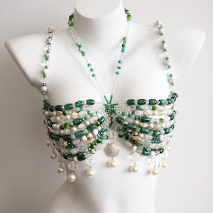 Könnte beinhalten: Ein dekoratives Bralette aus einer komplexen Anordnung von grünen, weißen und transparenten Perlen. Das Design besteht aus geschichteten Reihen ovaler und runder Perlen, akzentuiert durch hängende perlenartige Kugeln und einen zentralen Muschelanhänger.