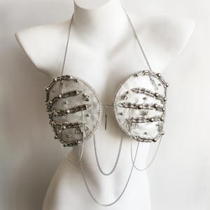 Può includere: Un reggiseno bianco stile allacciato al collo con decorazioni color argento, tra cui strutture a forma di costole, piccoli ciondoli a pipistrello e perline sferiche. Un ciondolo a spada argentata centrale unisce le coppe con catenelle.