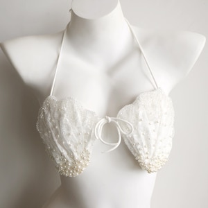 Könnte beinhalten: Ein weißes Neckholder-Bikini-Oberteil mit zwei muschelförmigen Cups, die mit filigraner weißer Spitze überzogen und mit zahlreichen kleinen weißen Perlen verziert sind. Das Kleidungsstück verfügt über dünne Nackenbänder und eine mittige Schnürung.