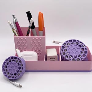 May include: A pink desk organizer with multiple compartments. It holds pens, markers, a charging block, and a pair of white earbuds. Two purple cable organizers are also included. The organizer has a honeycomb design.