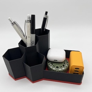 May include: Black desk organizer with red trim, holding pens, a white charging case, and a yellow power bank. The organizer has multiple compartments for office supplies and small electronics. The pens include a Sharpie and pens with the text "GRAND VELAS."