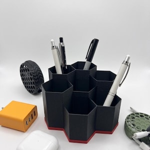 May include: A black and red hexagonal desk organizer holding pens. The organizer has a honeycomb design and is surrounded by other tech accessories, including a white charging case, a yellow charger, and a green cable organizer.