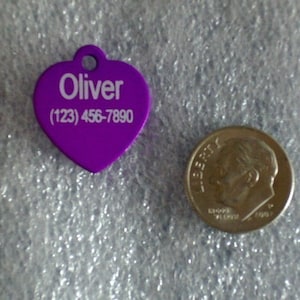 May include: Purple heart-shaped pet ID tag with white text that reads "Oliver (123) 456-7890".
