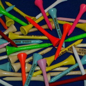 May include: A collection of colorful golf tees in various shades, including pink, green, yellow, and blue, with some featuring the text "Mike & Mona 6/16/2012". The tees are scattered on a blue surface.