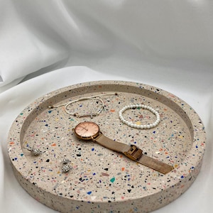 Könnte beinhalten: Ein runder, beigefarbener Terrazzoteller mit bunten Flecken hält eine roségoldene Uhr, Armbänder und Ohrstecker. Die Uhr hat ein Mesh-Armband. Der Teller ist ein stilvoller Organizer für Schmuck und Accessoires.