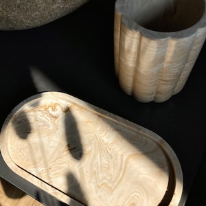 Peut inclure: Un plateau décoratif ovale avec un motif marbré beige et marron. Un vase cannelé de couleur claire est visible en arrière-plan. Les objets sont sur une surface sombre, avec des ombres.