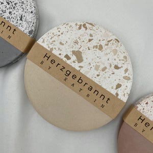 Könnte beinhalten: Eine Sammlung runder Untersetzer mit Terrazzo-Design. Jeder Untersetzer hat ein zweifarbiges Farbschema, mit einer hellbeigen unteren Hälfte und einer gesprenkelten oberen Hälfte. Die Untersetzer haben ein braunes Papierband mit dem Text "Herzgebrannt by Katrin".
