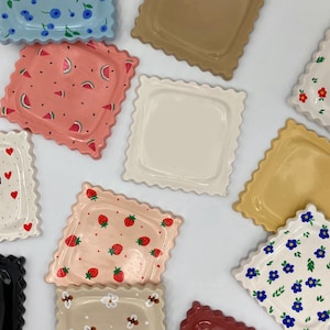 Peut inclure: Plusieurs assiettes carrées en céramique avec des bords festonnés. Les assiettes présentent divers motifs, notamment des myrtilles, des fraises, des tranches de pastèque et des motifs floraux. Les couleurs incluent le bleu clair, le rose, le beige et le blanc.