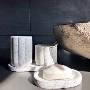 Könnte beinhalten: Ein Set aus gesprenkeltem, grauem Badezimmerzubehör. Das Set umfasst einen hohen Becher, ein kleines Gefäß mit Deckel und eine Seifenschale mit einem Stück Seife. Die Artikel bestehen aus einem strukturierten Material und stehen auf einer grauen Oberfläche.