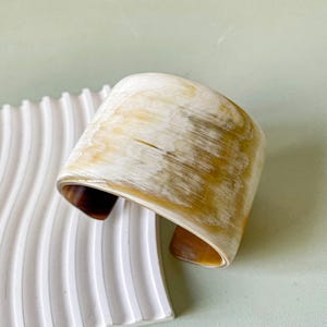 May include: A wide, open cuff bracelet with a natural, organic aesthetic. The bracelet features a blend of creamy white and golden-brown hues, with a polished finish. The cuff is approximately 2 inches wide.