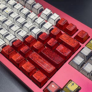 Puede incluir: Un teclado mecánico rojo con un esquema de color rojo y negro. El teclado tiene una variedad de teclas, incluidas teclas de función, teclas numéricas y teclas de letras. Las teclas son todas rojas y tienen una superficie texturizada.