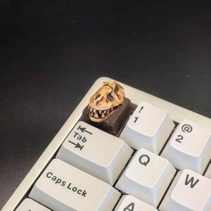 May include: A custom keycap featuring a detailed dinosaur skull design in shades of brown and tan. The keycap is mounted on a keyboard with white keys, including the Tab, Caps Lock, and number keys.