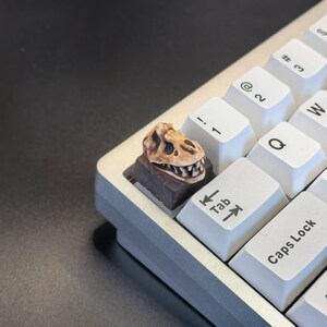 May include: A custom keyboard with a dinosaur skull keycap. The keycap is a detailed, tan-colored Tyrannosaurus Rex skull on a dark brown base. The keyboard has white keys and a silver frame.
