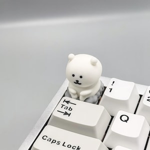 May include: A white keyboard with a small white bear figurine sitting on the Tab key. The bear has black eyes and a small black nose.