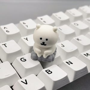 May include: A white bear-shaped keycap sits on a white keyboard. The keycap is sitting on the 'G' key. The keyboard has black lettering on the keys.