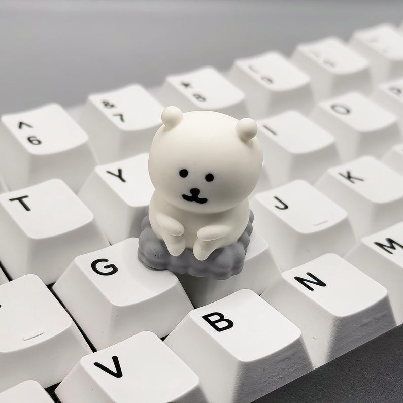 May include: A white bear-shaped keycap sits on a white keyboard. The keycap is sitting on the 'G' key. The keyboard has black lettering on the keys.