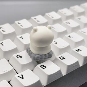 May include: A white keycap with a gray cloud design. The keycap is shaped like a cartoon character with a round head and a small body. The keycap is sitting on a white keyboard.