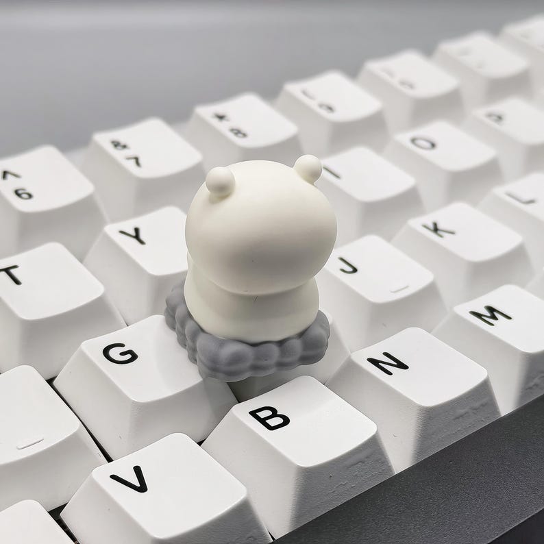 May include: A white keycap with a gray cloud design. The keycap is shaped like a cartoon character with a round head and a small body. The keycap is sitting on a white keyboard.