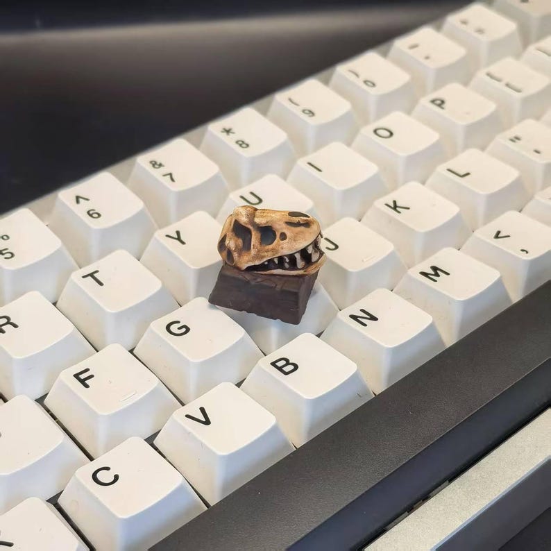 May include: A custom keyboard keycap featuring a detailed dinosaur skull design. The keycap is brown and tan, mounted on a dark brown base, and sits atop a white keyboard. The keyboard has white keycaps with black lettering.